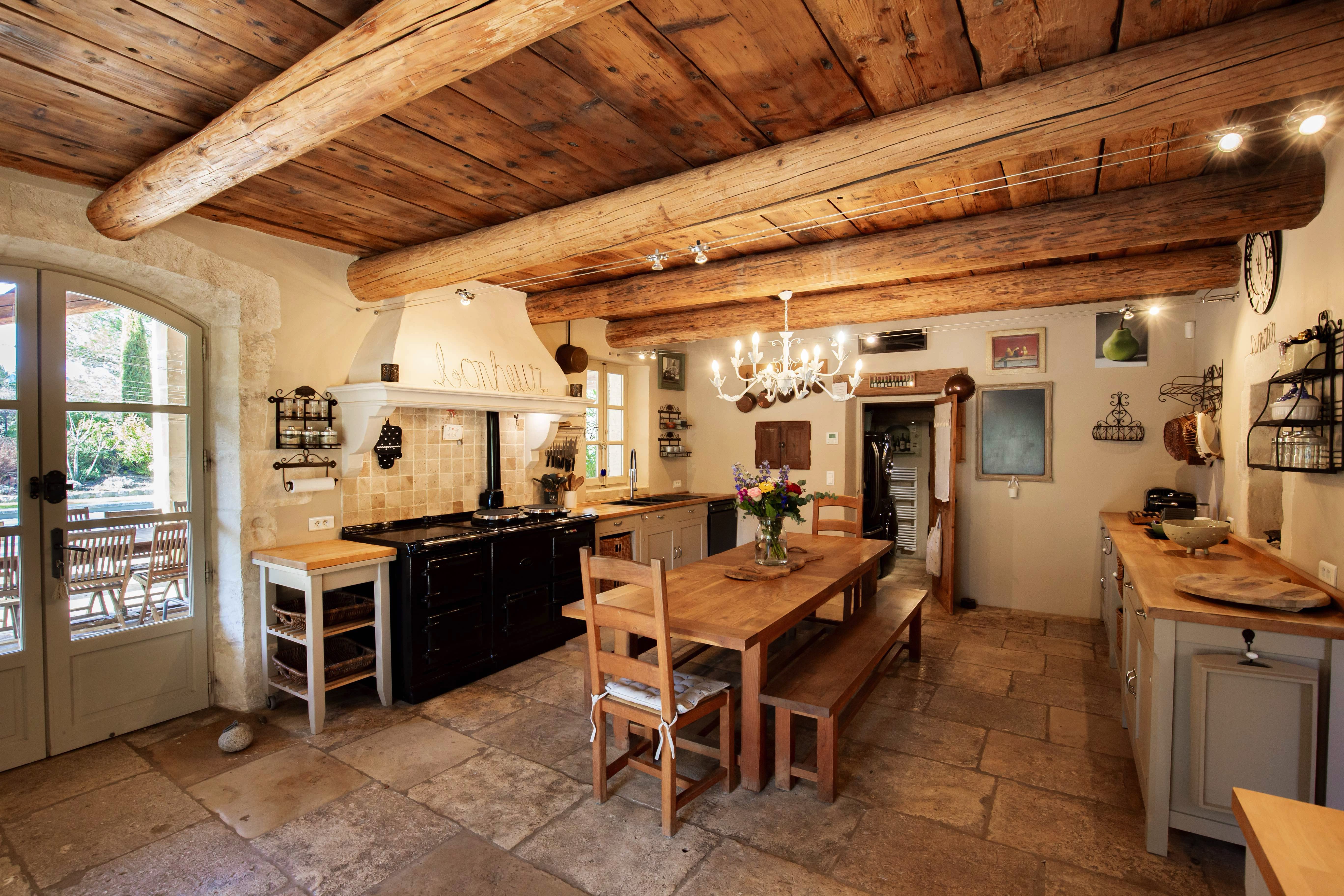 Spacious kitchen with rustic charm, featuring exposed wooden beams, stone flooring, a large central island, wooden dining table, and modern appliances. the room is well-lit with natural light from windows.